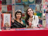 Two women at an anime convention in San José, Costa Rica, posing with peace signs, surrounded by art and comics.