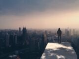 A person stands on a rooftop, gazing over a foggy city skyline at dusk.