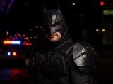 Batman cosplayer in detailed costume at a night event with police lights in the background.