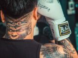 A boxer with intricate tattoos and boxing gloves gets ready for training indoors.